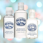 Three bottles of Boy Butter "Clear" Water-Based Lubricant are showcased against a blurred background. The lubricant is made in the USA, variably sized, and labeled as safe for use with condoms.